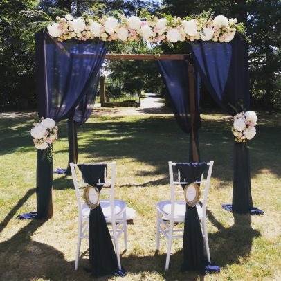 Arche de cérémonie avec grosses compositions florales et chaises des mariés avec voilage bleu marine